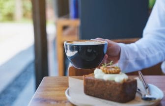 Closeup image of a woman holding and drinking coffee with a piece of carrot cake on the table | コーヒーステーション
