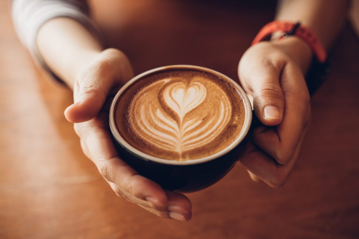 Close up Cup of coffee latte in coffee shopFemale hands holding a cup of coffee cup with heart shaped latte art foam on black wood table | コーヒーステーション