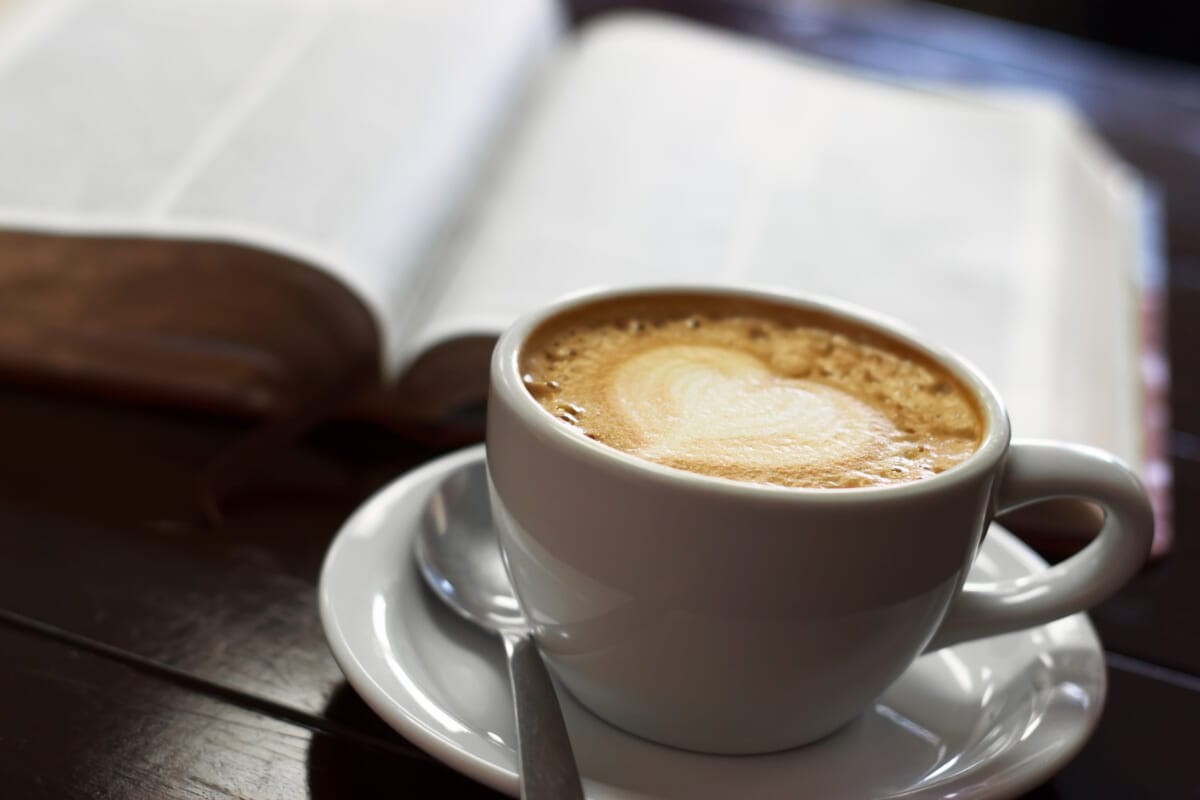 A cup of latte coffee on a tabletop next to a christian bible in spanish | コーヒーステーション