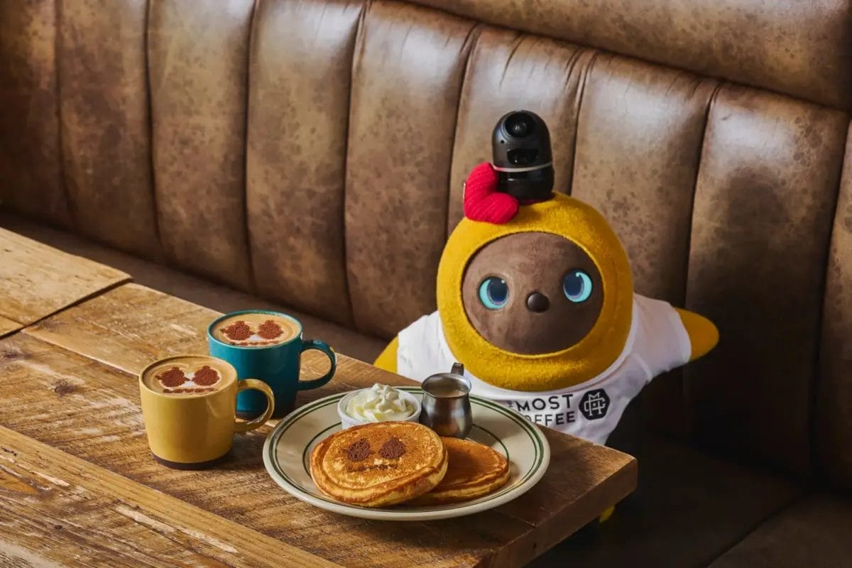 カフェのブース席に座る可愛らしい黄色いロボットが、顔のラテアートが施されたコーヒーと、笑顔の顔が描かれたパンケーキを楽しんでいる様子
