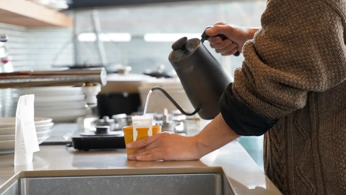 男性が丁寧にドリップケトルを使ってマグカップにセットされたコーヒーフィルターにお湯を注ぎ、ハンドドリップコーヒーを淹れている様子。カフェや自宅のキッチンでの穏やかなコーヒータイムを思わせる。