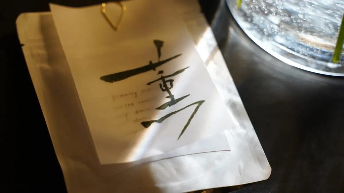 白いパッケージに墨で「重」と書かれた和紙のラベルが金色のクリップで留められています。背景には水泡が輝くグラスと緑の茎が見え、光が差し込む静かで上品な雰囲気の画像です。