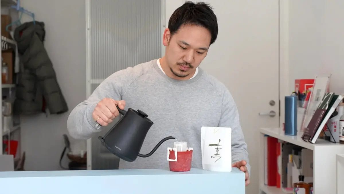 男性が黒い電気ケトルを使ってドリップコーヒーを淹れている様子を捉えた画像です。手元には白いコーヒーの袋と赤いカップがあり、丁寧にコーヒーを準備している雰囲気が伝わります。