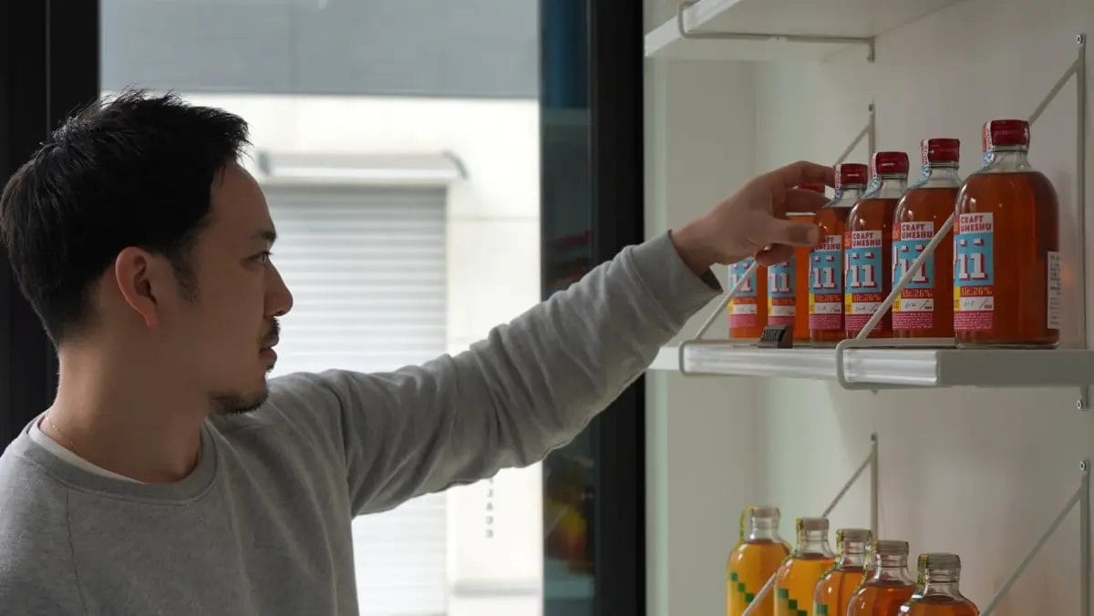 一人の男性が白い棚に並べられた「CRAFT UMESHU」と書かれたボトルに手を伸ばしている様子。店内のような空間で、オレンジ色の液体が入ったボトルが複数陳列されている。